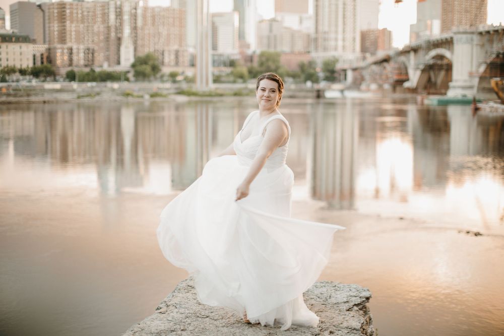 Bride spinning white gown on rocks by river with Minneapolis skyline at golden hour.