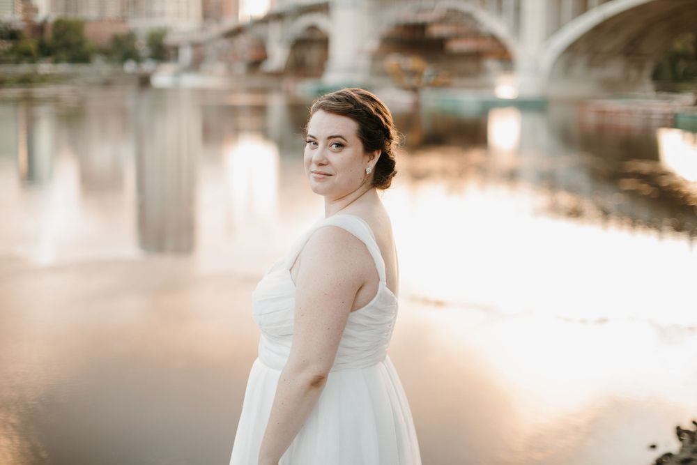 Bride in white gown posing by river with bridge and city skyline in soft golden light.