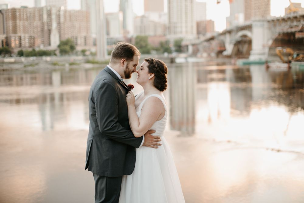 Bride and groom share a romantic kiss by the river with a city skyline reflecting on the water behind them.