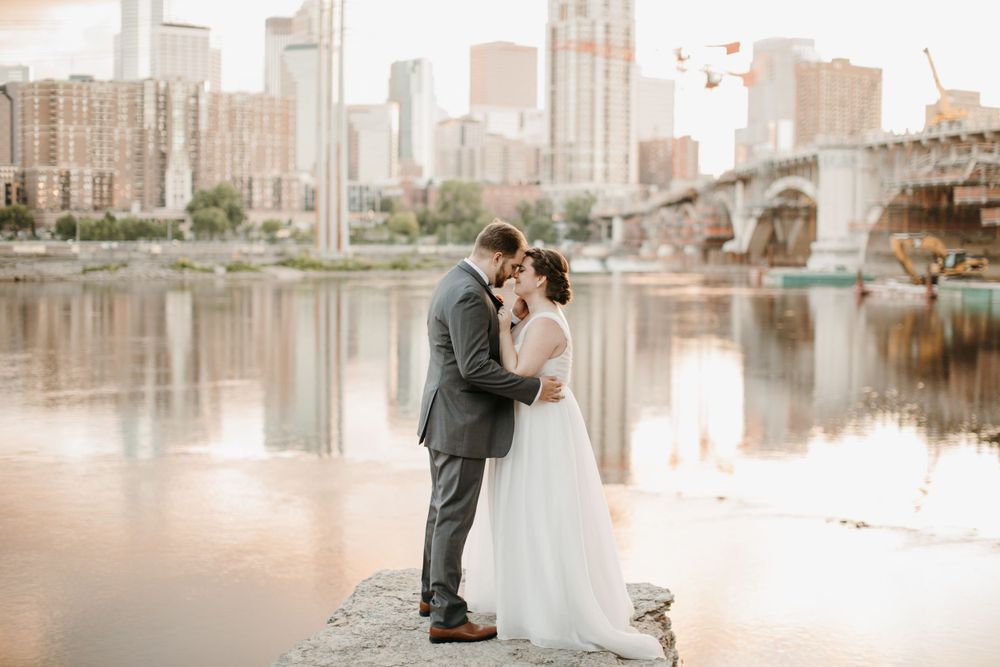 Wedding couple embraces on riverside rocks with a stunning city skyline and bridge reflected in calm waters.