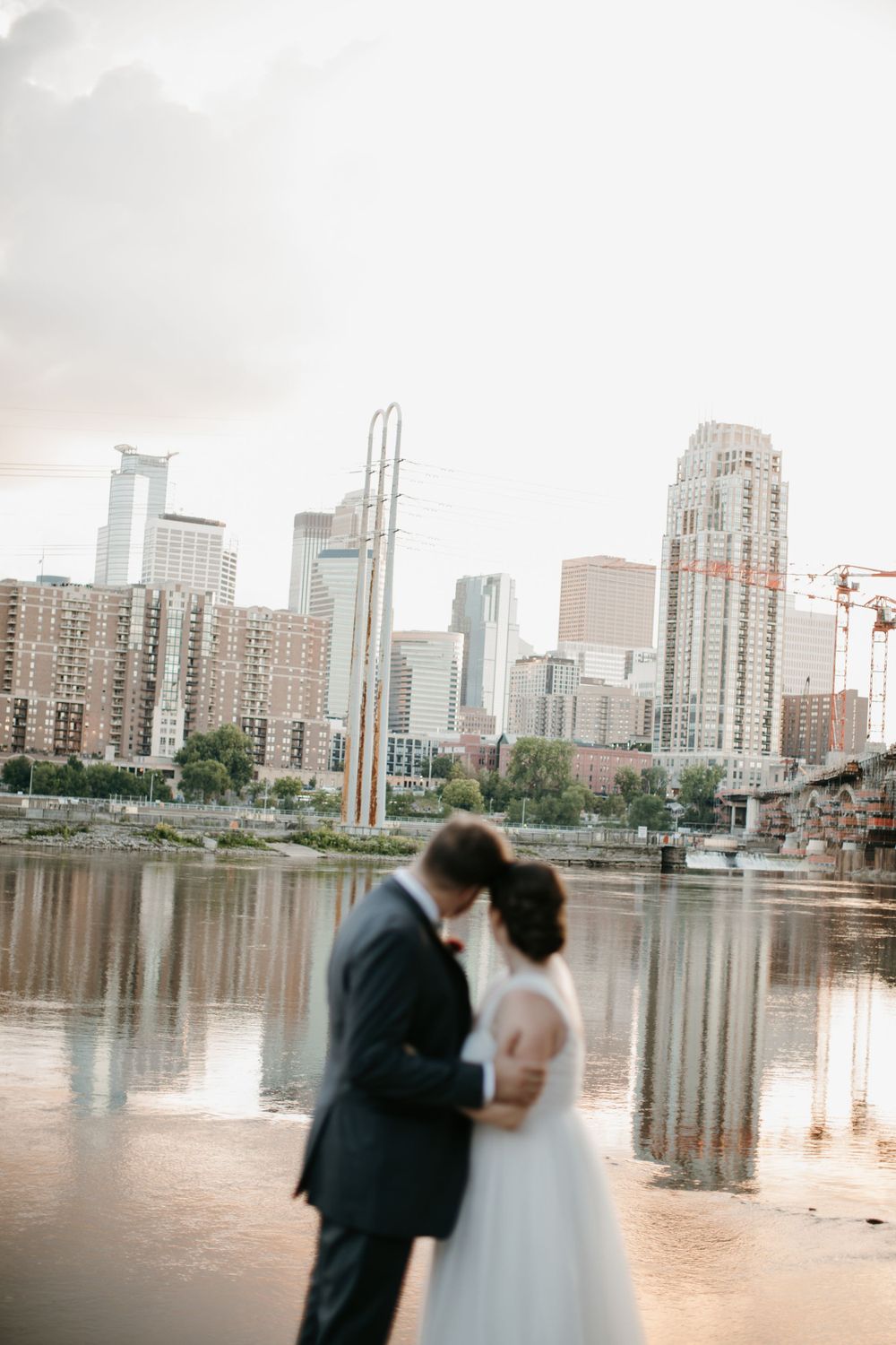 Bride and groom share an intimate moment by the river with a dramatic urban skyline backdrop at golden hour.