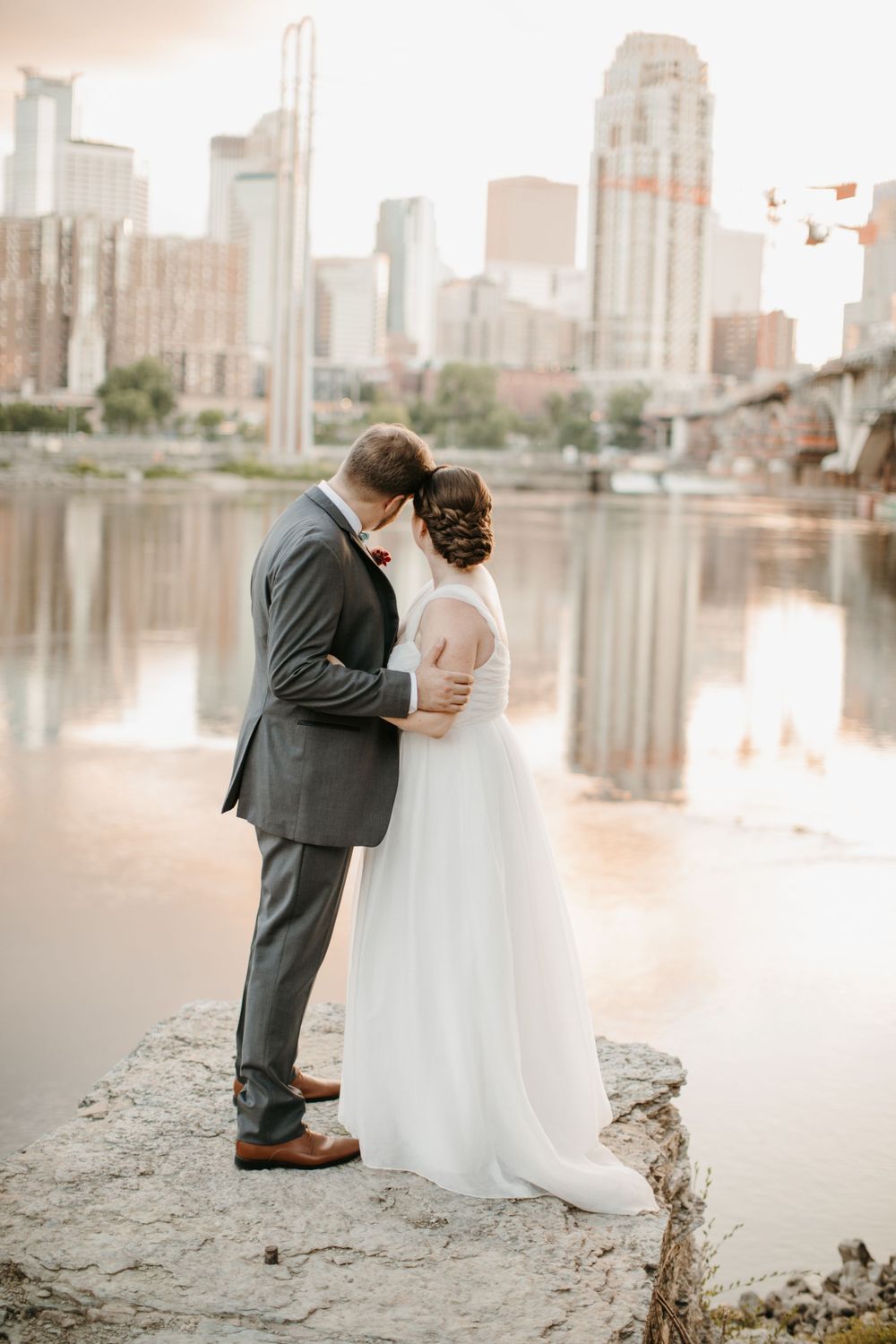 Bride and groom embrace tenderly on rocky riverbank with a glowing city skyline reflected in the water.