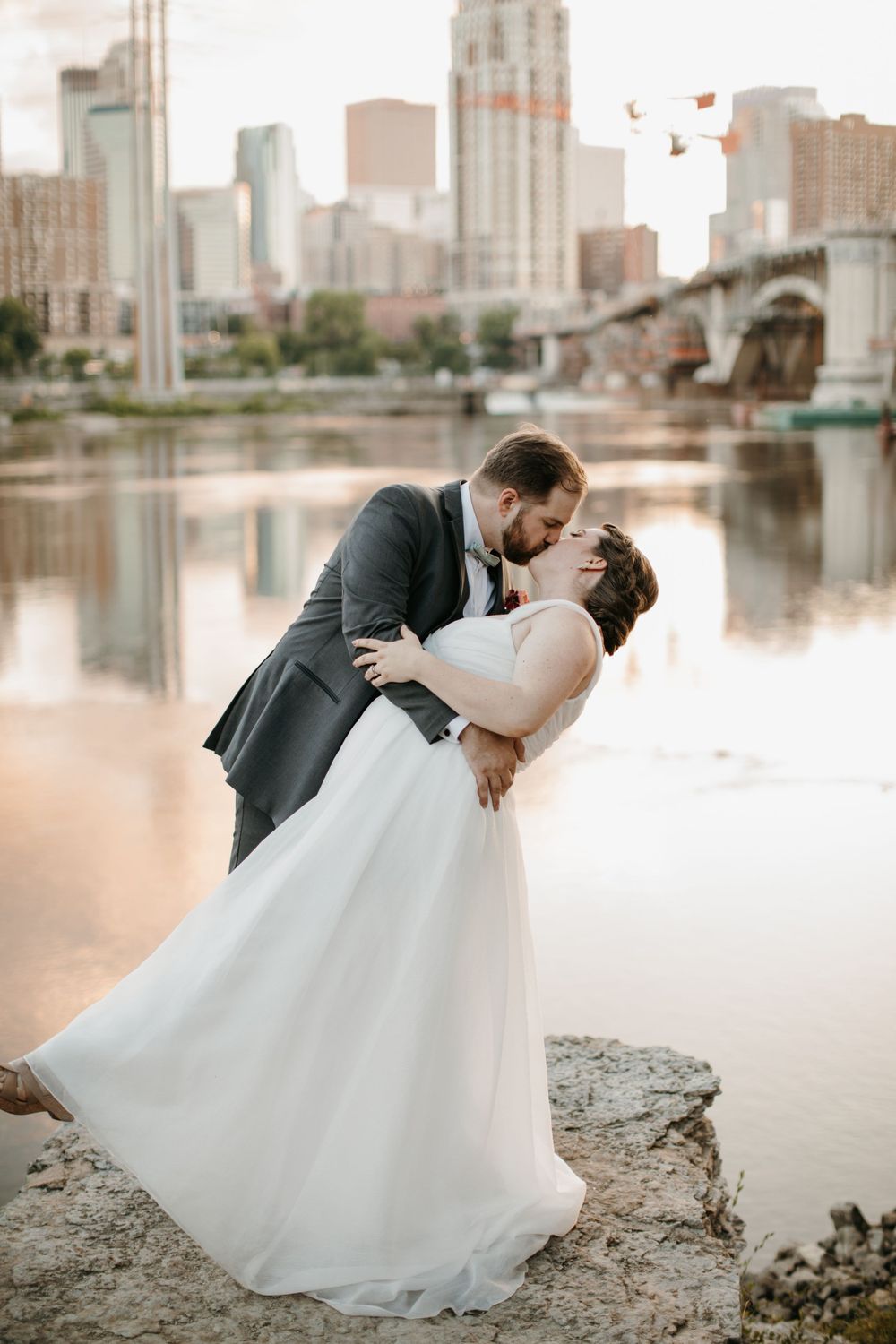 Groom dips bride for a passionate kiss on rocky riverbank with city skyline and bridge in the background.