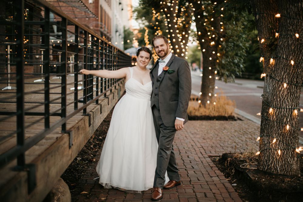 Smiling bride and groom pose together on a brick sidewalk with trees wrapped in glowing fairy lights.