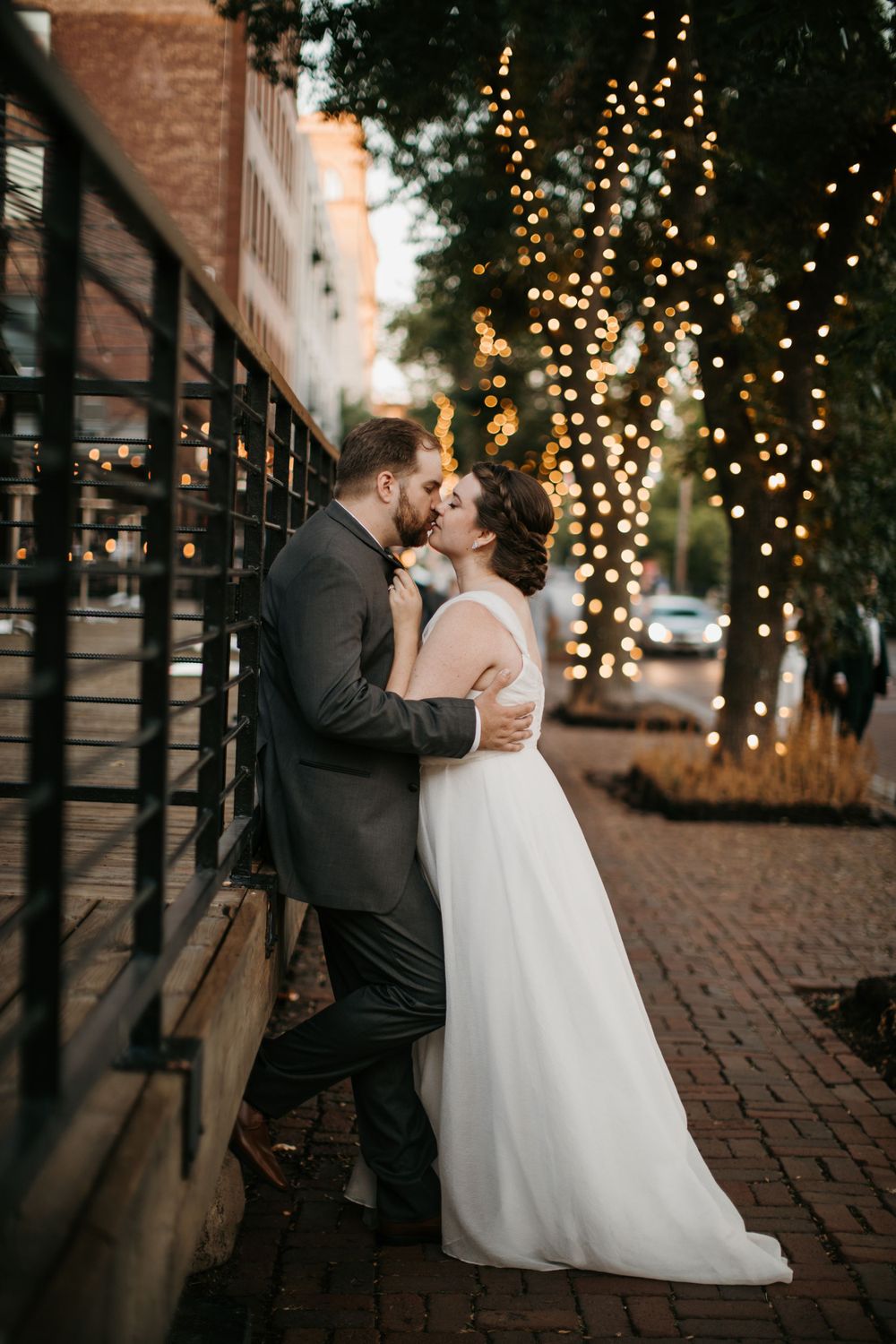 Bride and groom share a romantic kiss against a wall on a brick street with twinkling fairy lights.