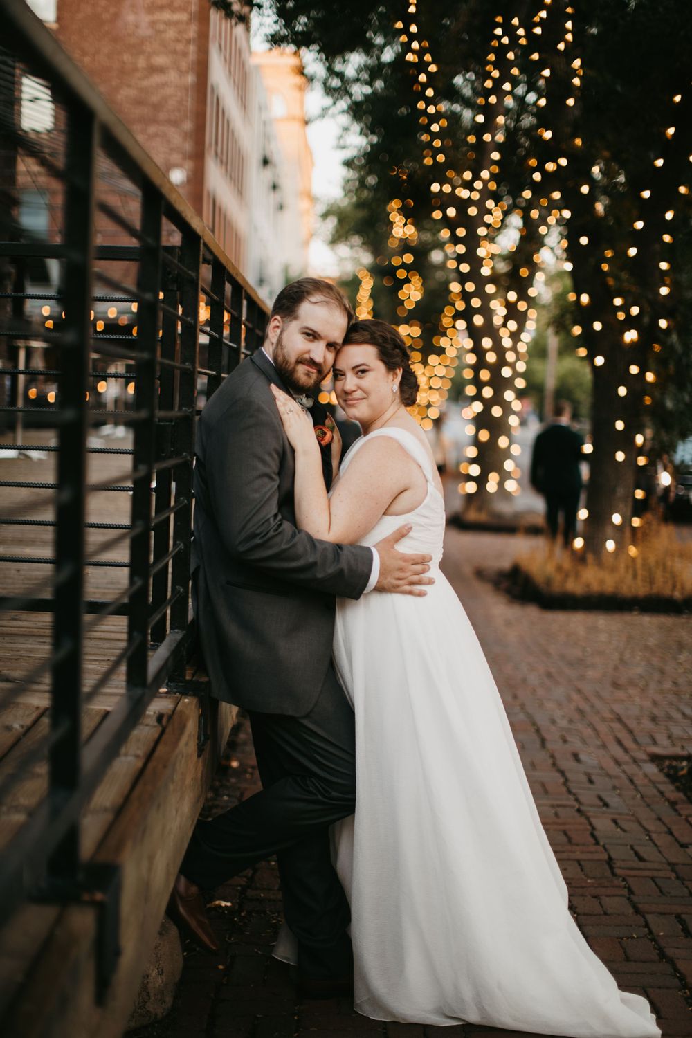 Bride and groom embrace against a railing on a brick street with bokeh fairy lights glowing behind them.