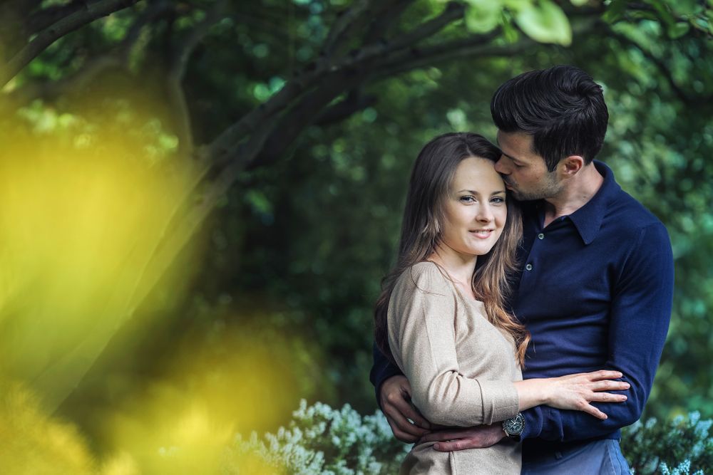 Newly engaged male kisses his fiancées forehead under the shade of branches in Walkden Gardens