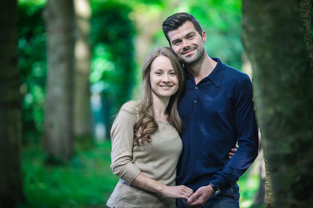 Engaged smiling couple stand close together surrounded by green leaves and trees at Walkden Gardens in Sale