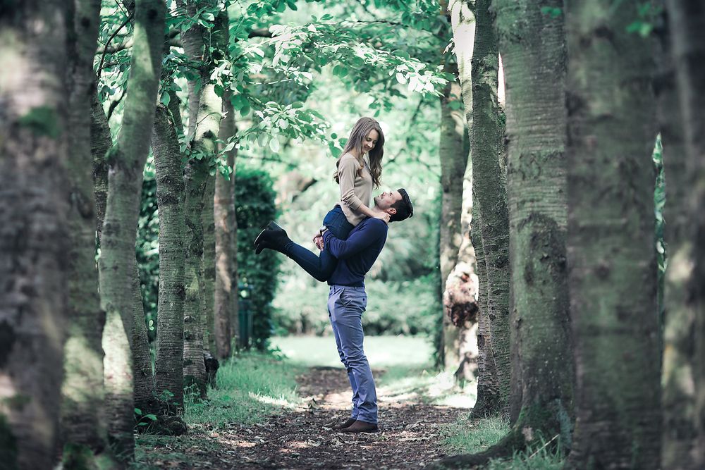 Man playfully lifts his fiancée up in his arms between a row of trees in Walkden Park Manchester
