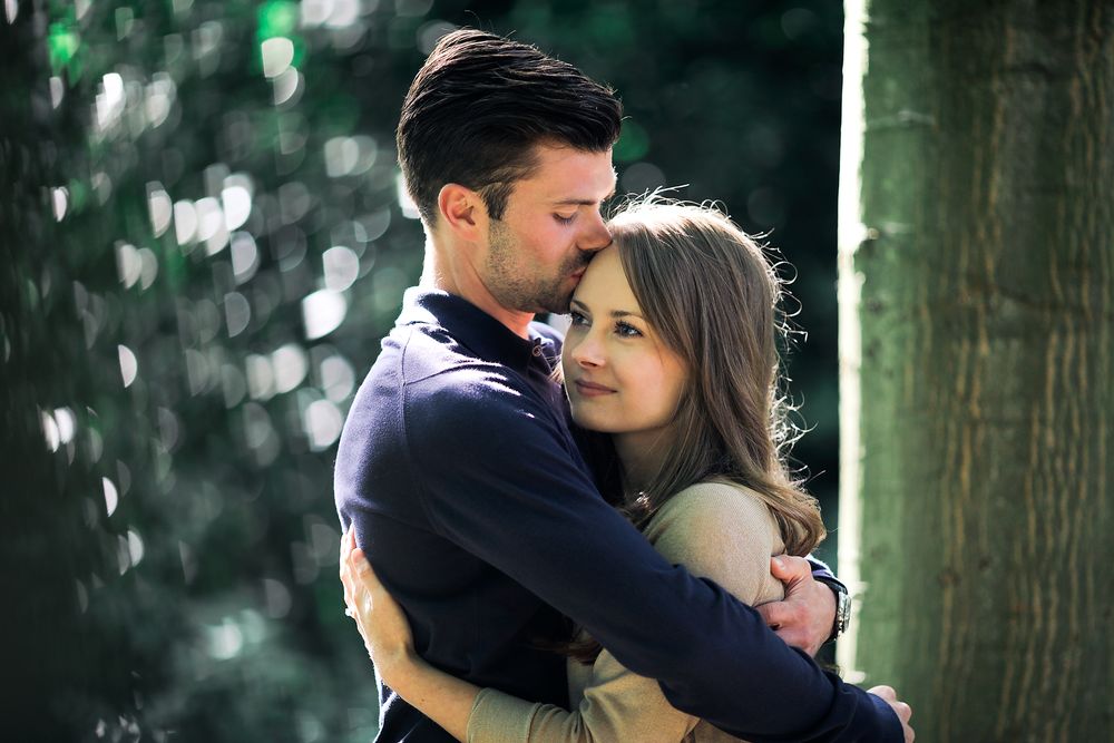 Engaged man and woman fondly hold each other in the sunlight against a tree trunk in the park