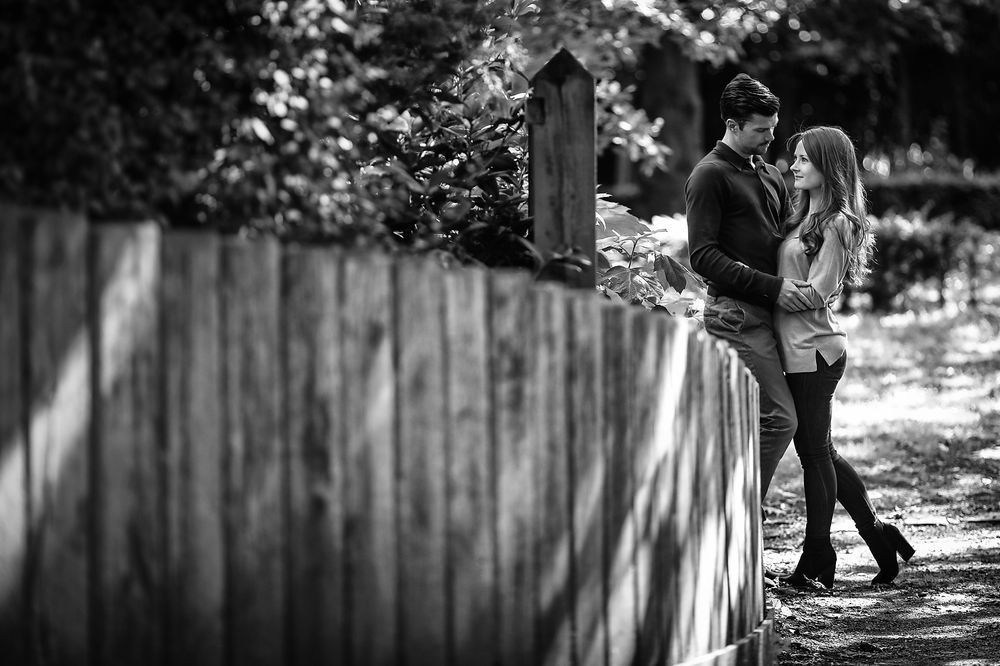 Young man and woman cuddle standing next to a long wooden fence in the park during summer