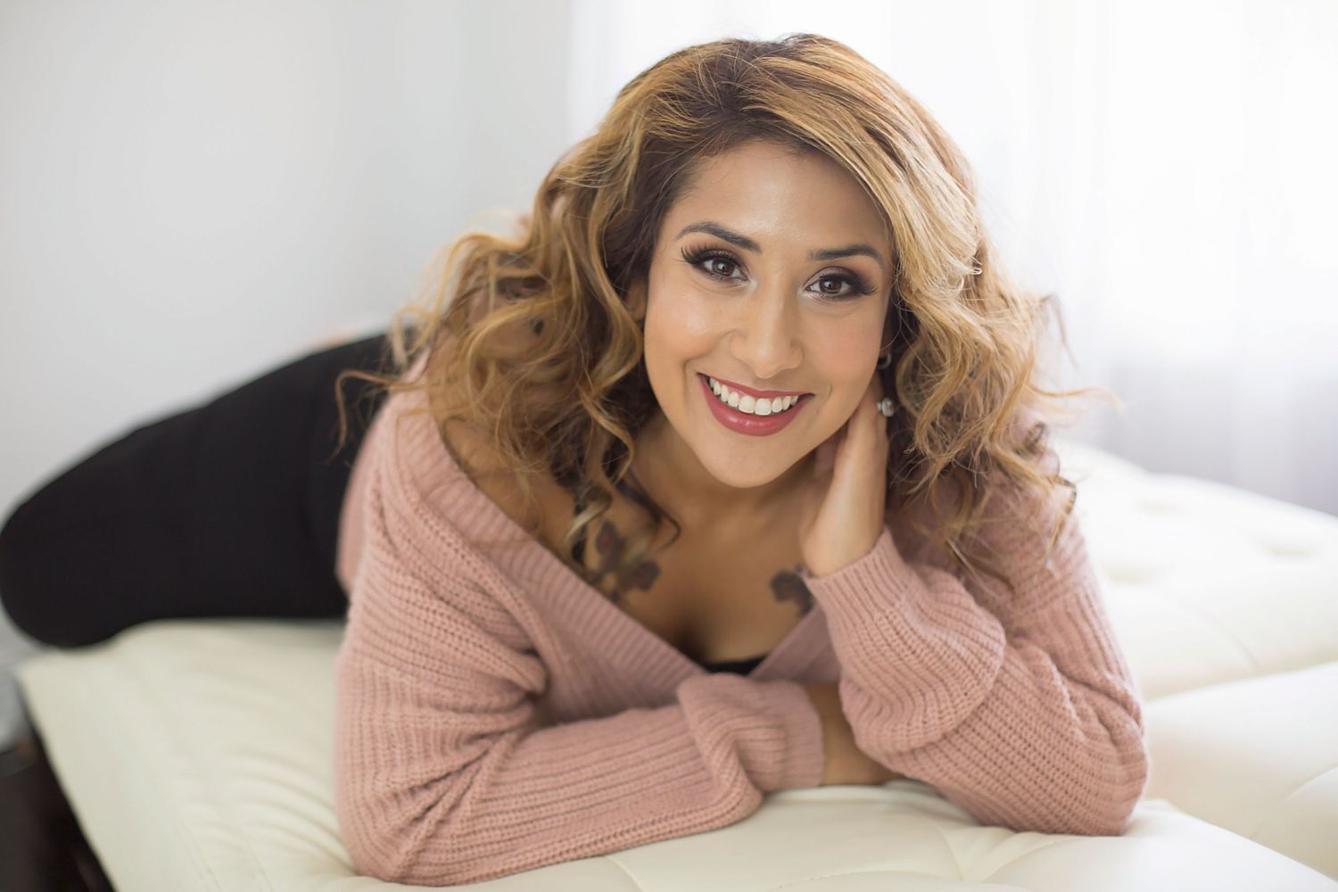 A cheerful woman with curly blonde hair wearing a pink sweater relaxes on a white bed smiling.