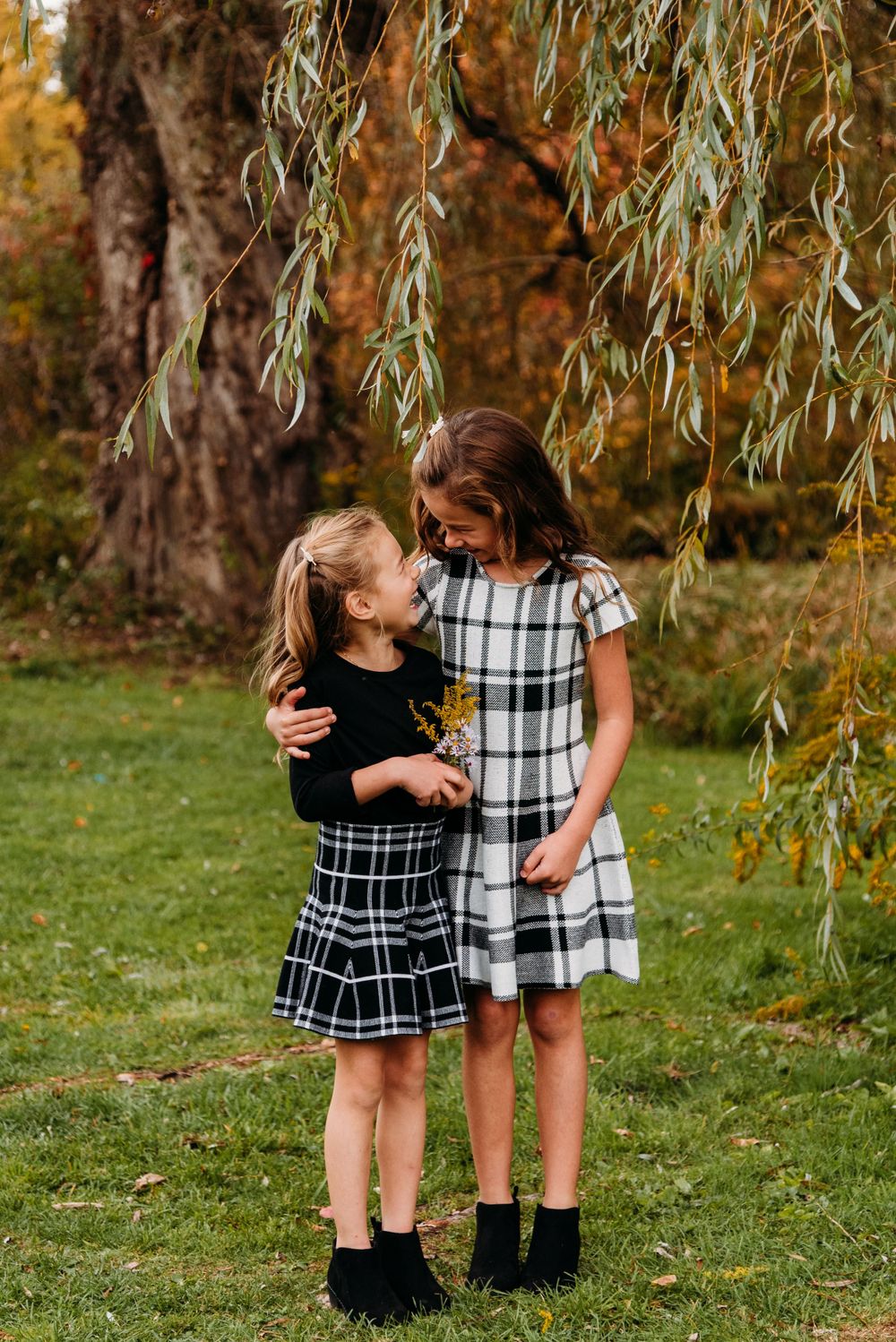 Two children in matching plaid dresses share a moment under weeping willow tree.