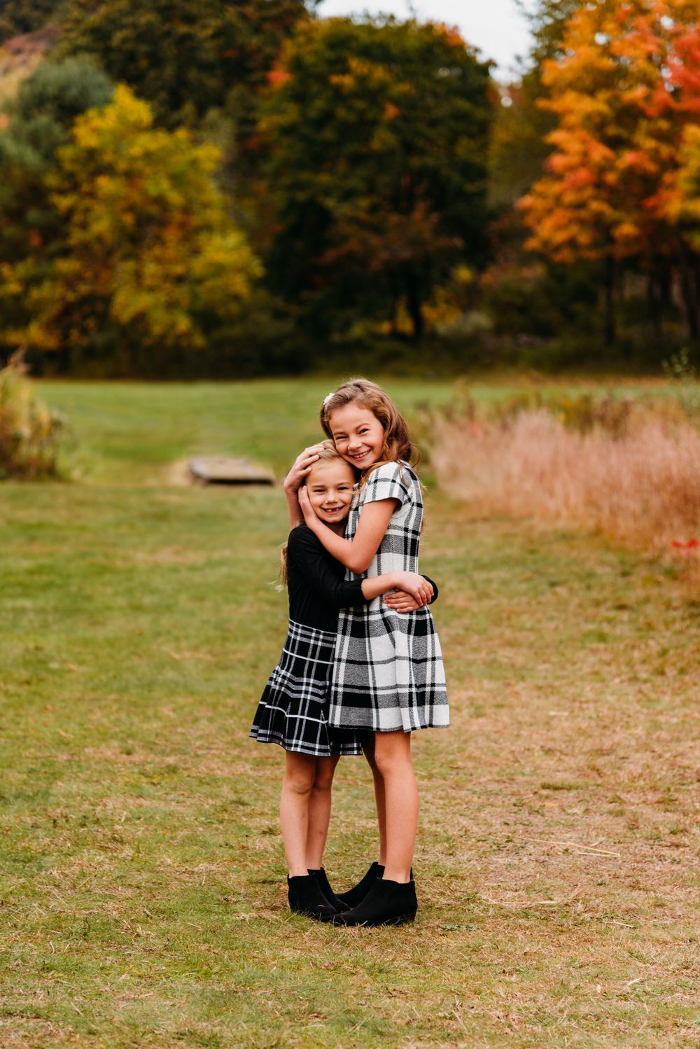 Two children in plaid dresses hugging and smiling outdoors in autumn setting.