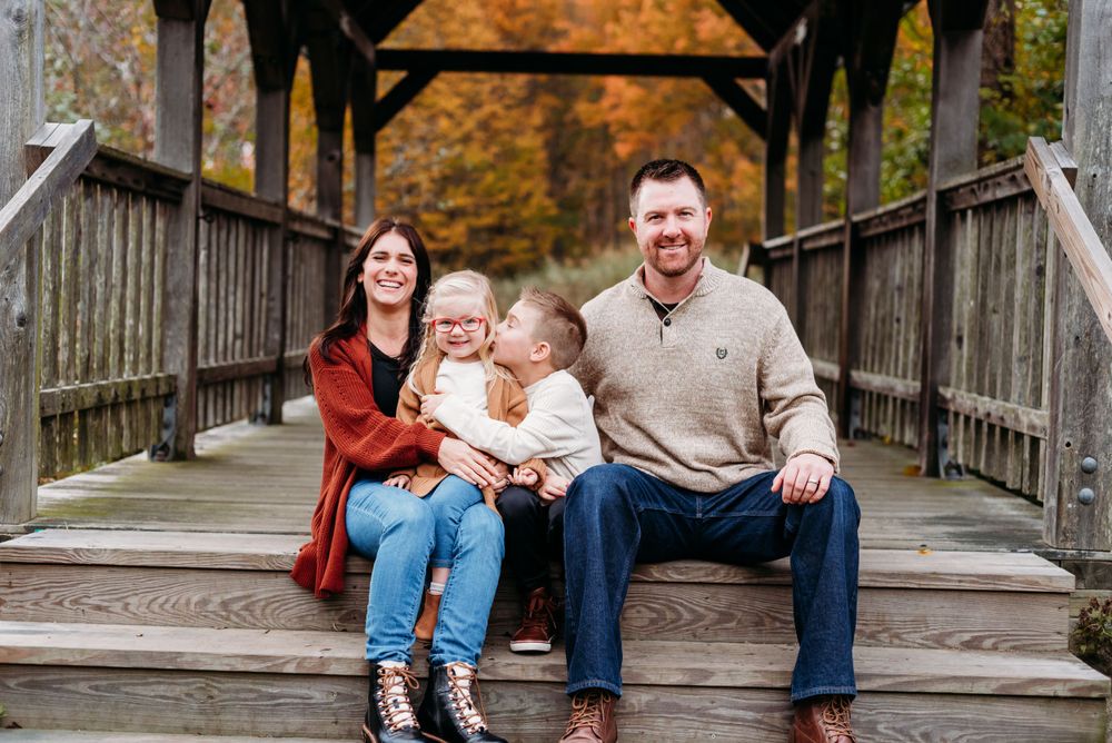 A family sits together on a wooden covered bridge during autumn with colorful fall foliage in the background.