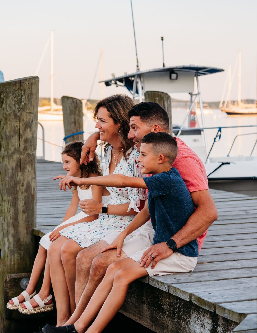 A family enjoys quality time sitting together on a dock surrounded by boats at sunset.