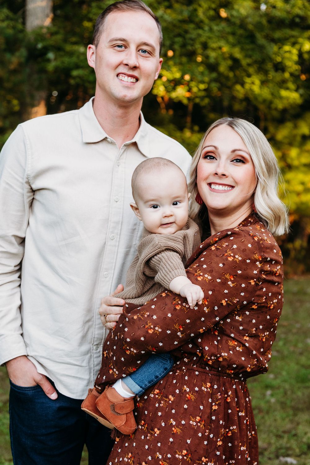 Outdoor family portrait with baby in casual attire against autumn foliage background.