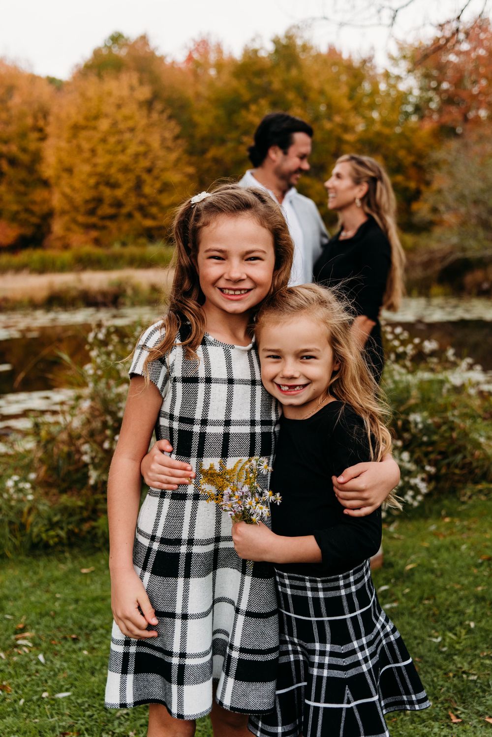 Two young girls in plaid dresses embrace while standing in an autumn garden with fall foliage.