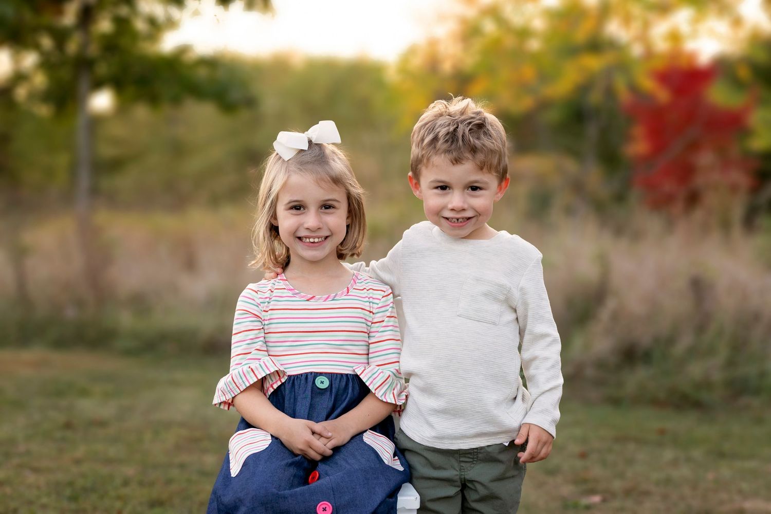 Two children stand together outdoors with fall foliage in background at sunset.
