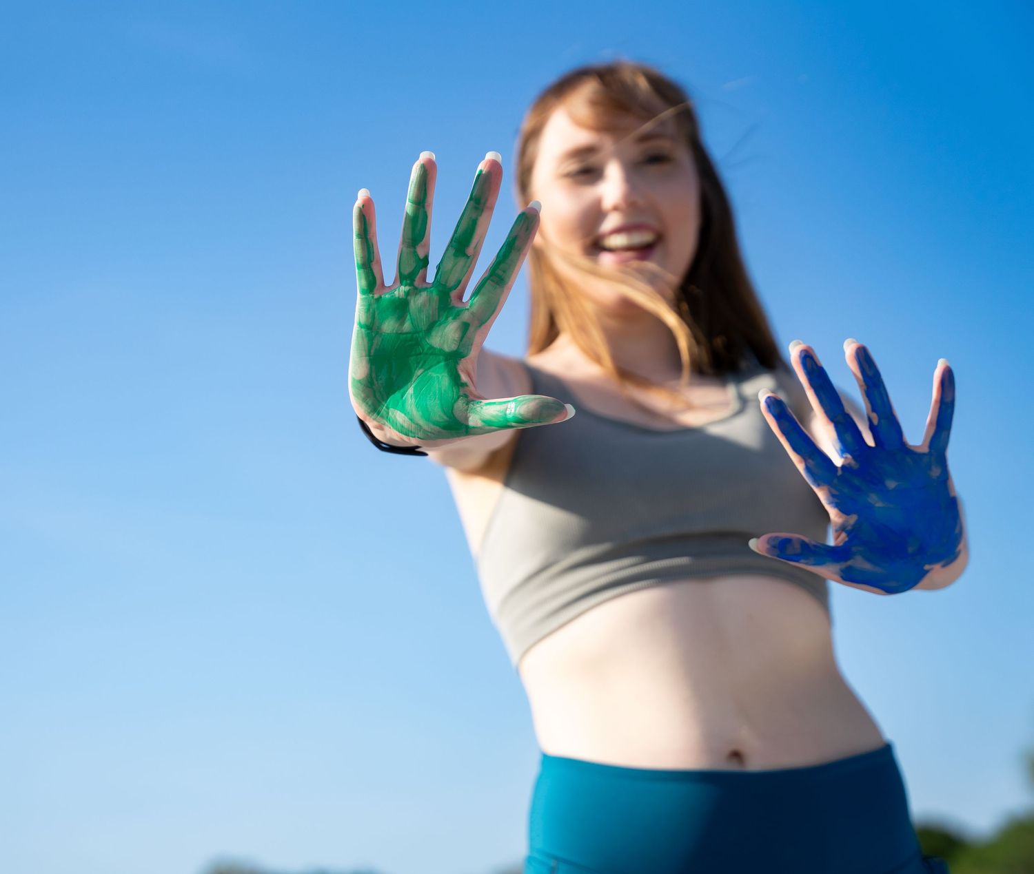 Hands covered in bright blue and green paint held up against a clear blue sky.