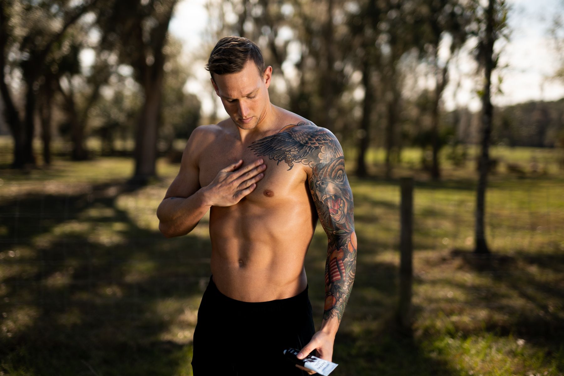 Shirtless person with tattoos standing in a sunlit park during golden hour.