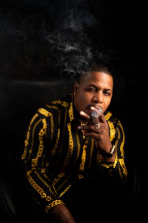Person wearing patterned gold and black jacket smoking in dramatic lighting.