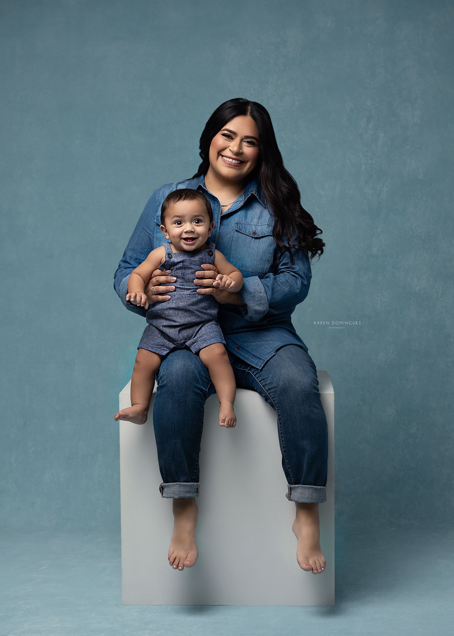 Parent and child in matching denim outfits sit casually on white blocks against blue backdrop.
