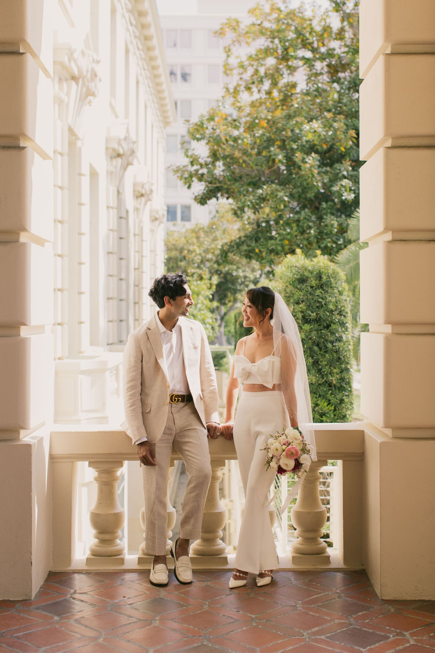 A couple in wedding attire shares a romantic moment on an elegant columned balcony with lush greenery.