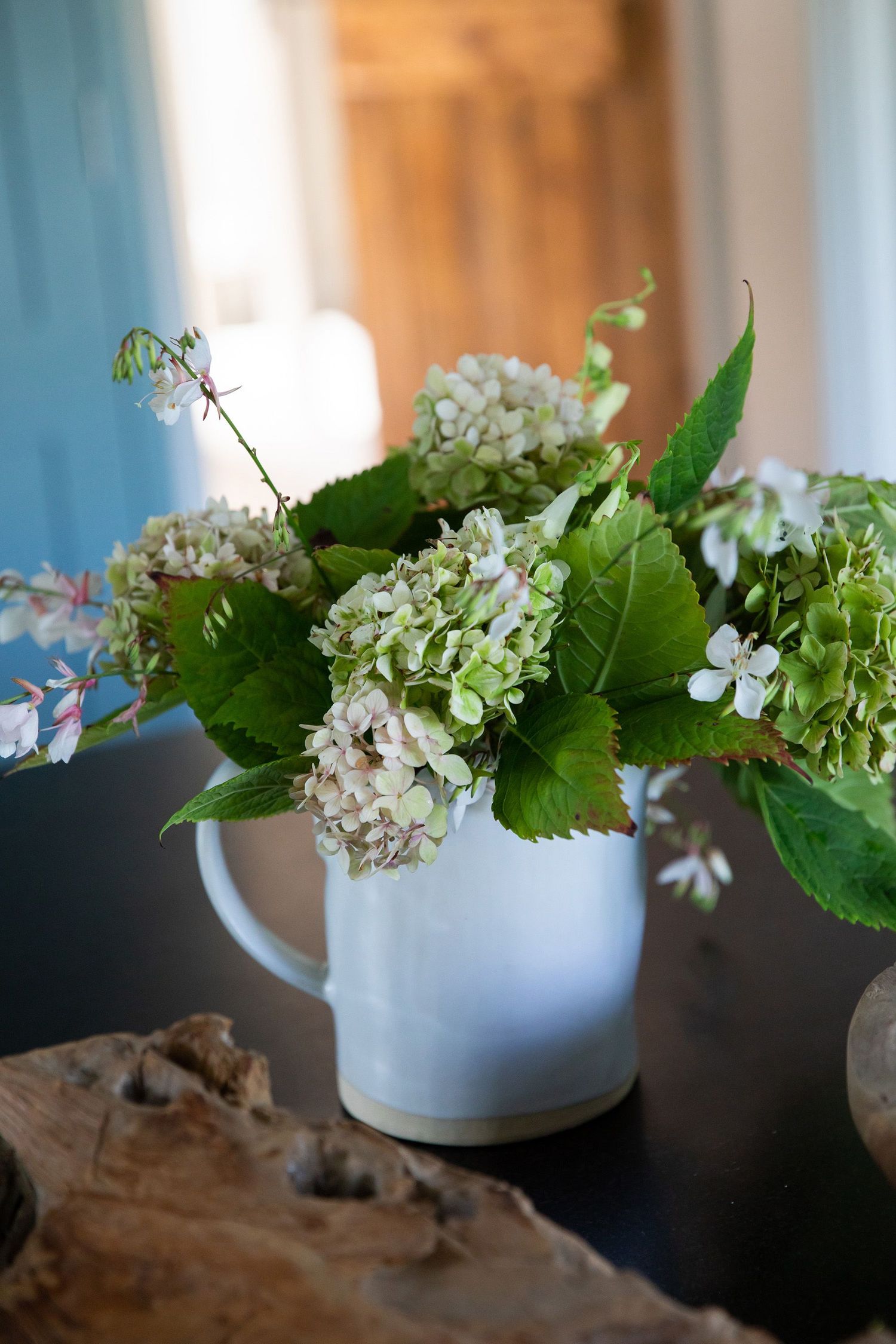 Fresh white hydrangea flowers arranged in a ceramic pitcher on a wooden surface with natural light.