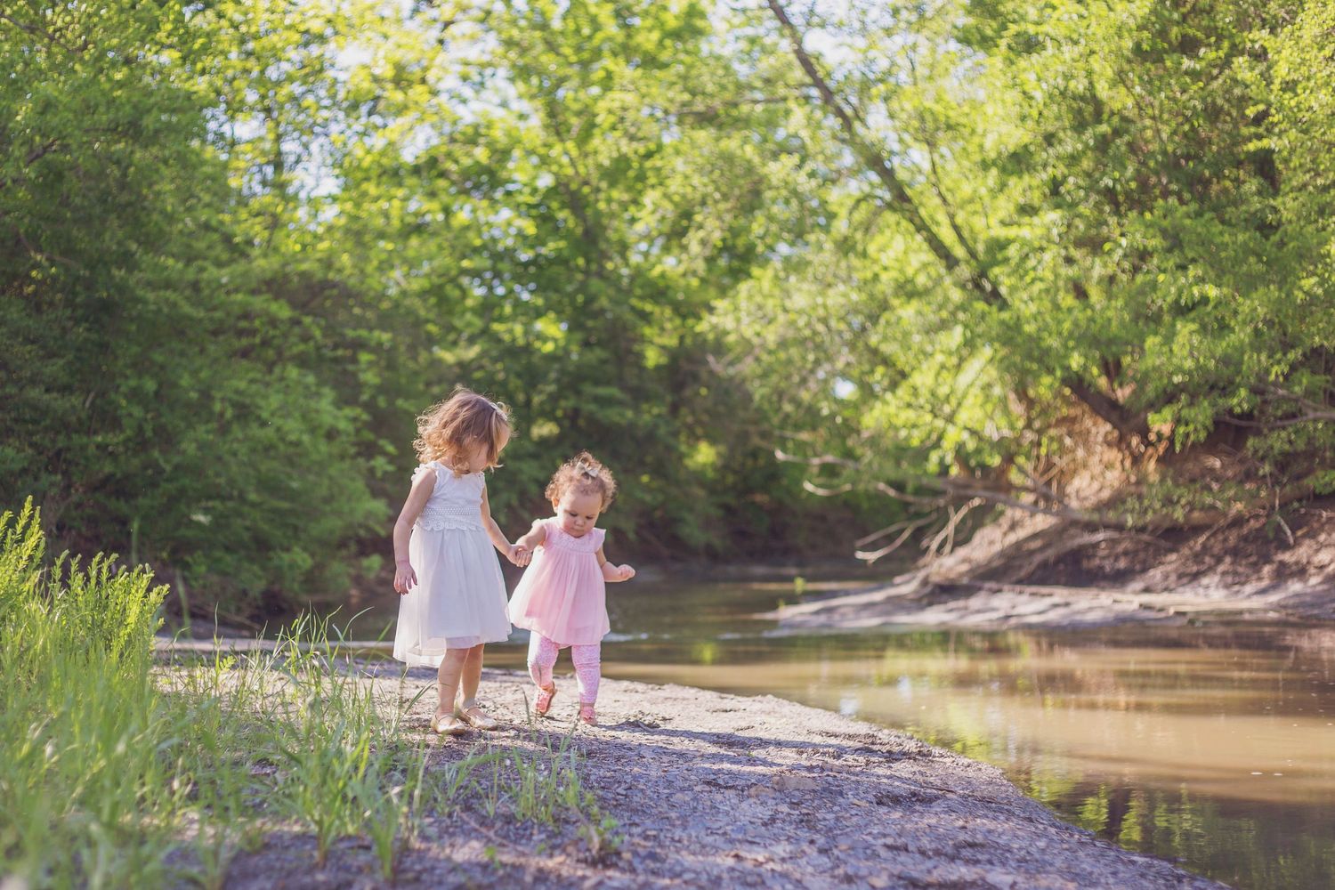 Two children in white dresses explore a peaceful creek bank on a sunny summer day.