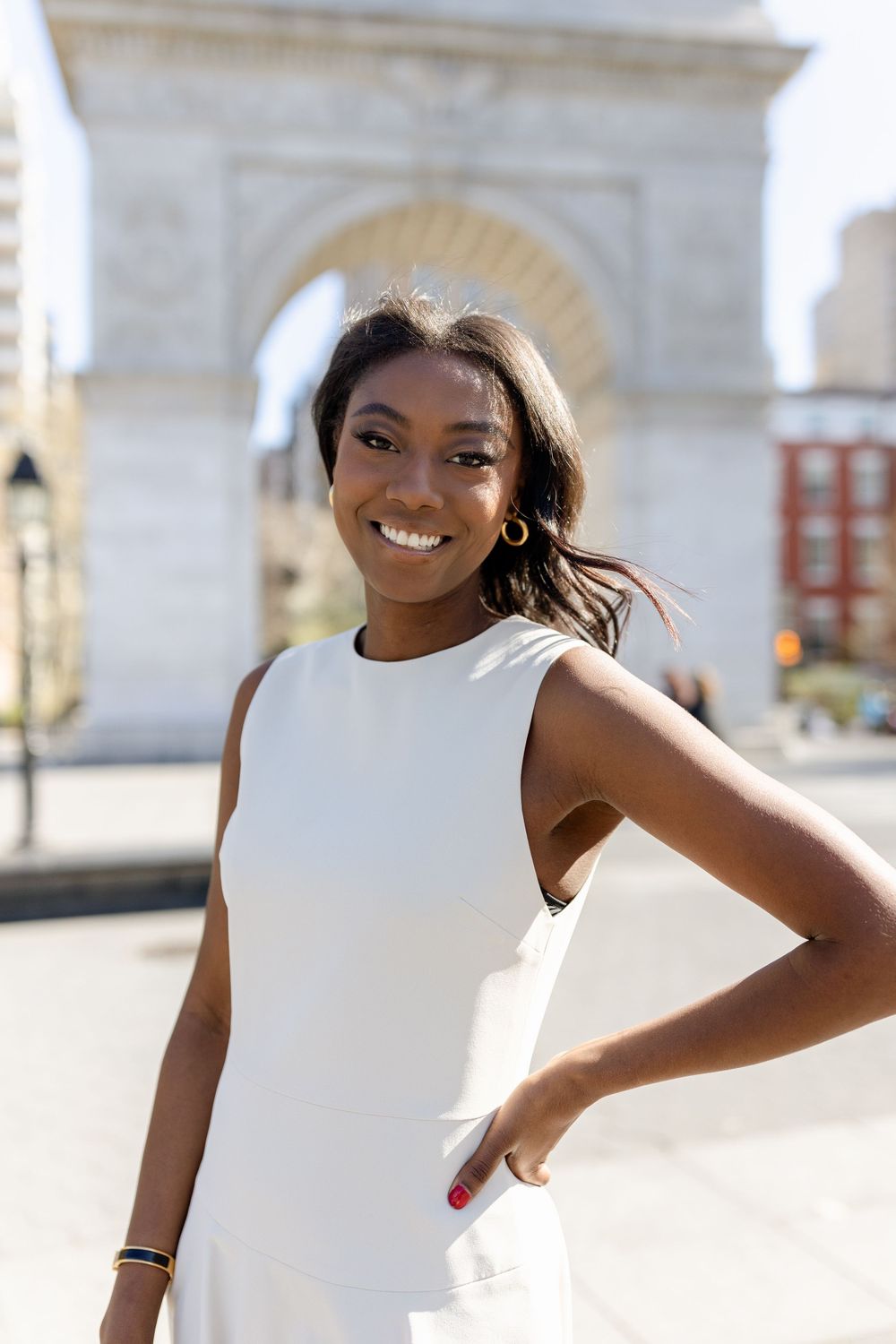 Student in sleeveless white dress poses confidently near iconic NYU arch.
