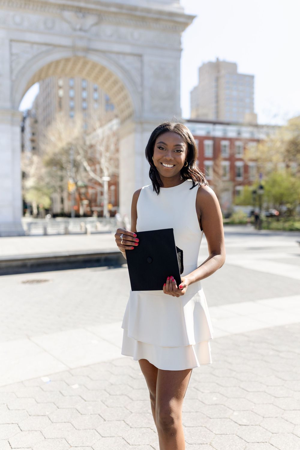 Graduate in white dress poses with portfolio near Washington Square Arch.