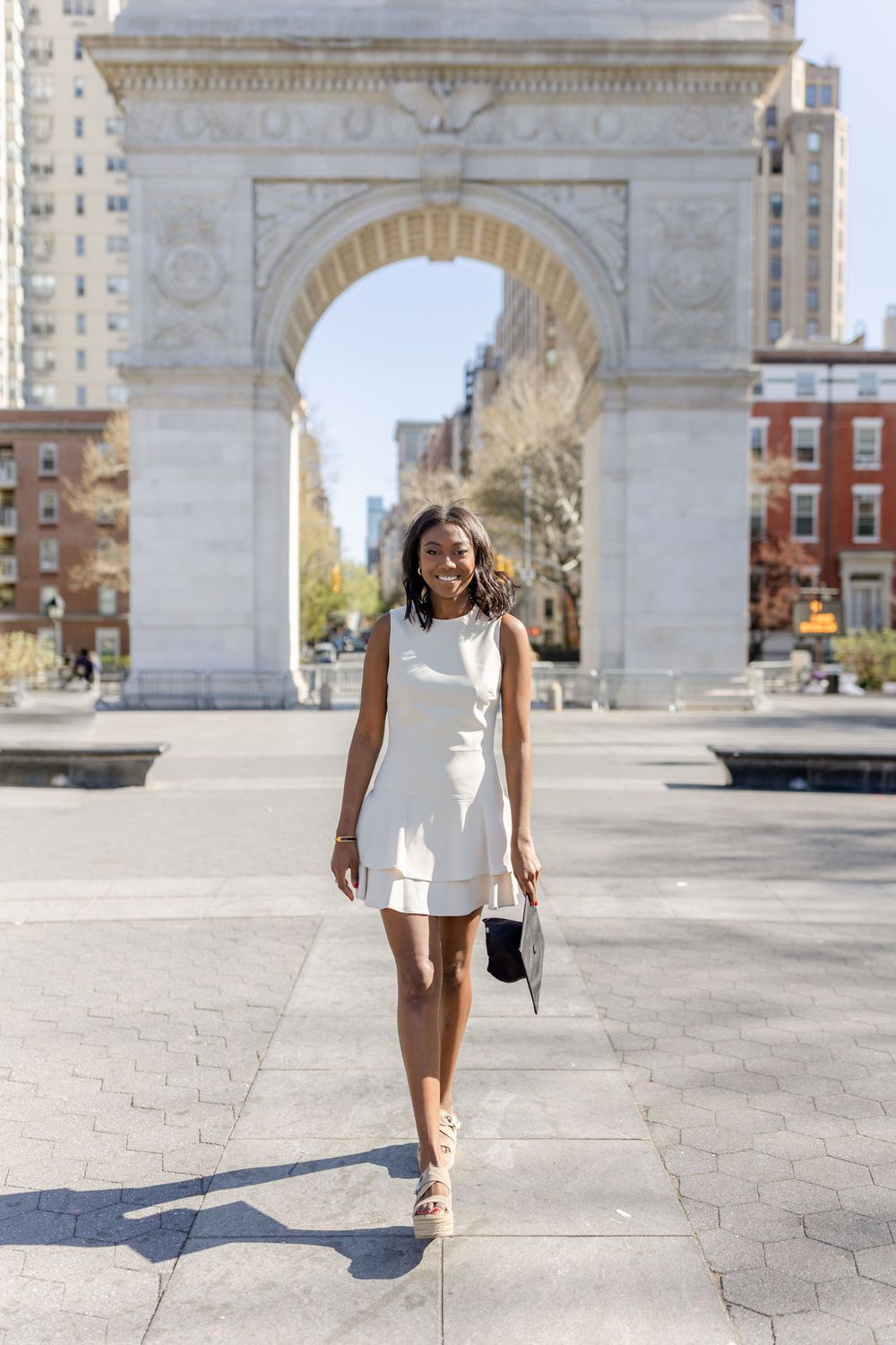 Graduate in white dress walks beneath Washington Square Arch on sunny day.