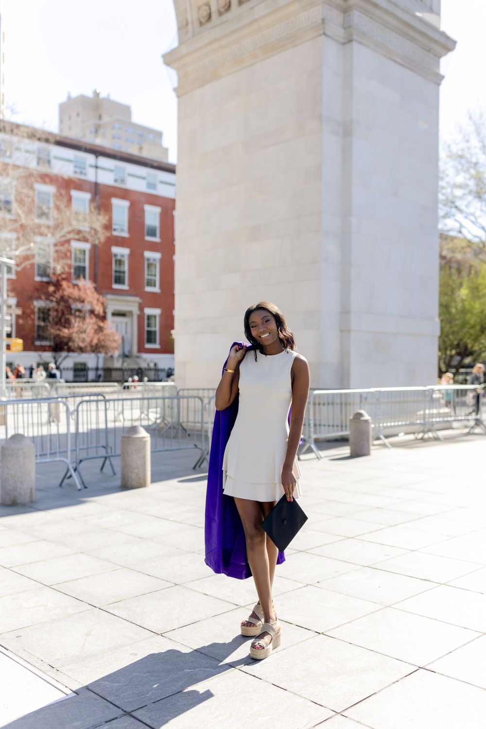 Student carrying graduation gown poses in white dress near Washington Square Arch.