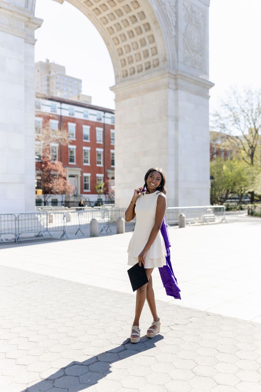 Graduate in white dress poses under Washington Square Arch holding diploma and purple graduation gown.