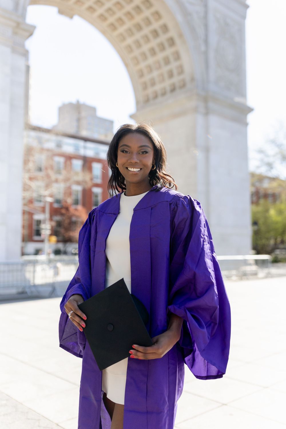Graduate wearing purple graduation regalia and white dress stands proudly by historic arch.