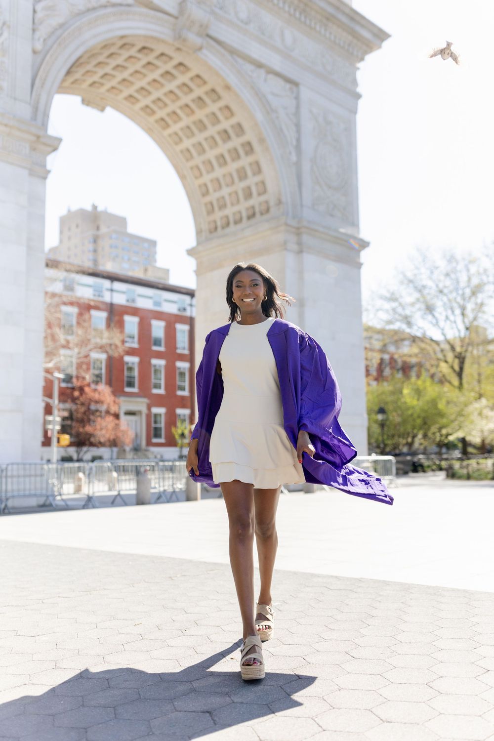 Graduate in flowing purple gown and white dress walks confidently under marble arch.