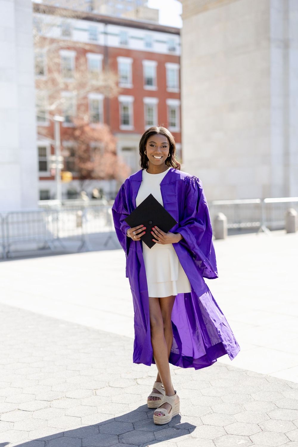Graduate holds diploma while posing in purple regalia under Washington Square Arch.