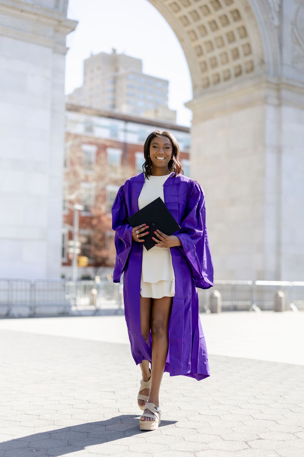Graduate walks elegantly in purple graduation gown and white dress under marble archway.