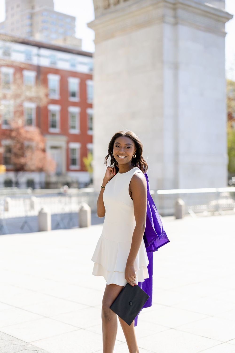 Graduate in sleeveless white dress poses with purple gown and diploma by historic arch.