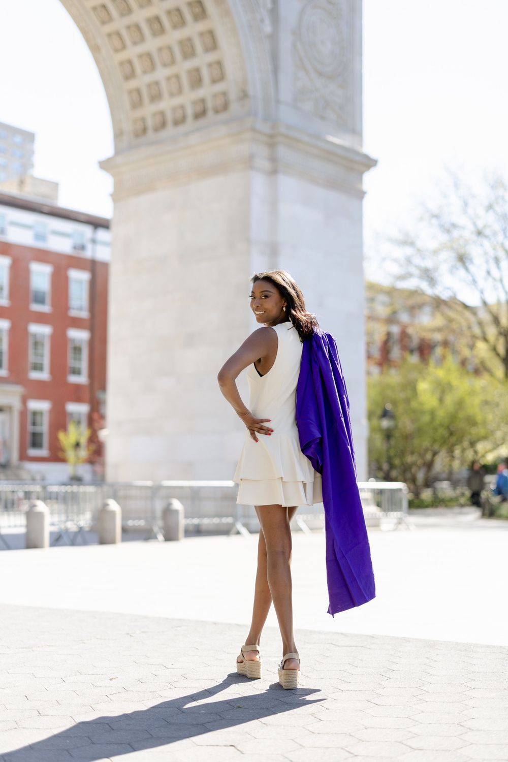 Graduate wearing white dress and purple academic gown poses gracefully in front of Washington Square Arch in NYC.