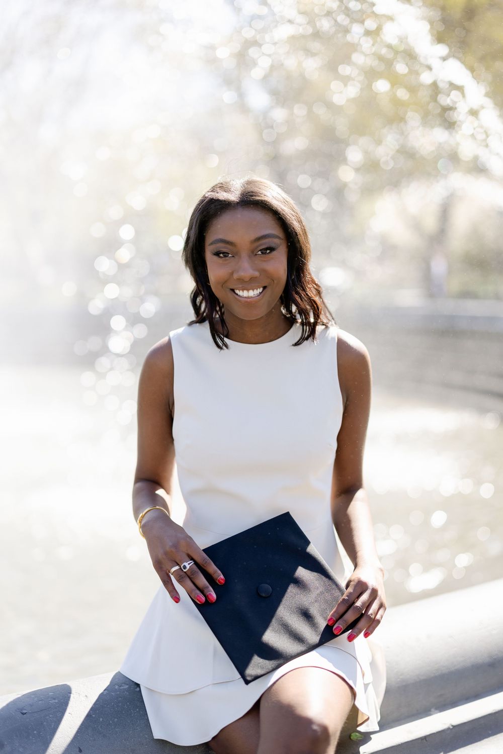 Graduate in white dress holds diploma while sitting outdoors with bokeh background.