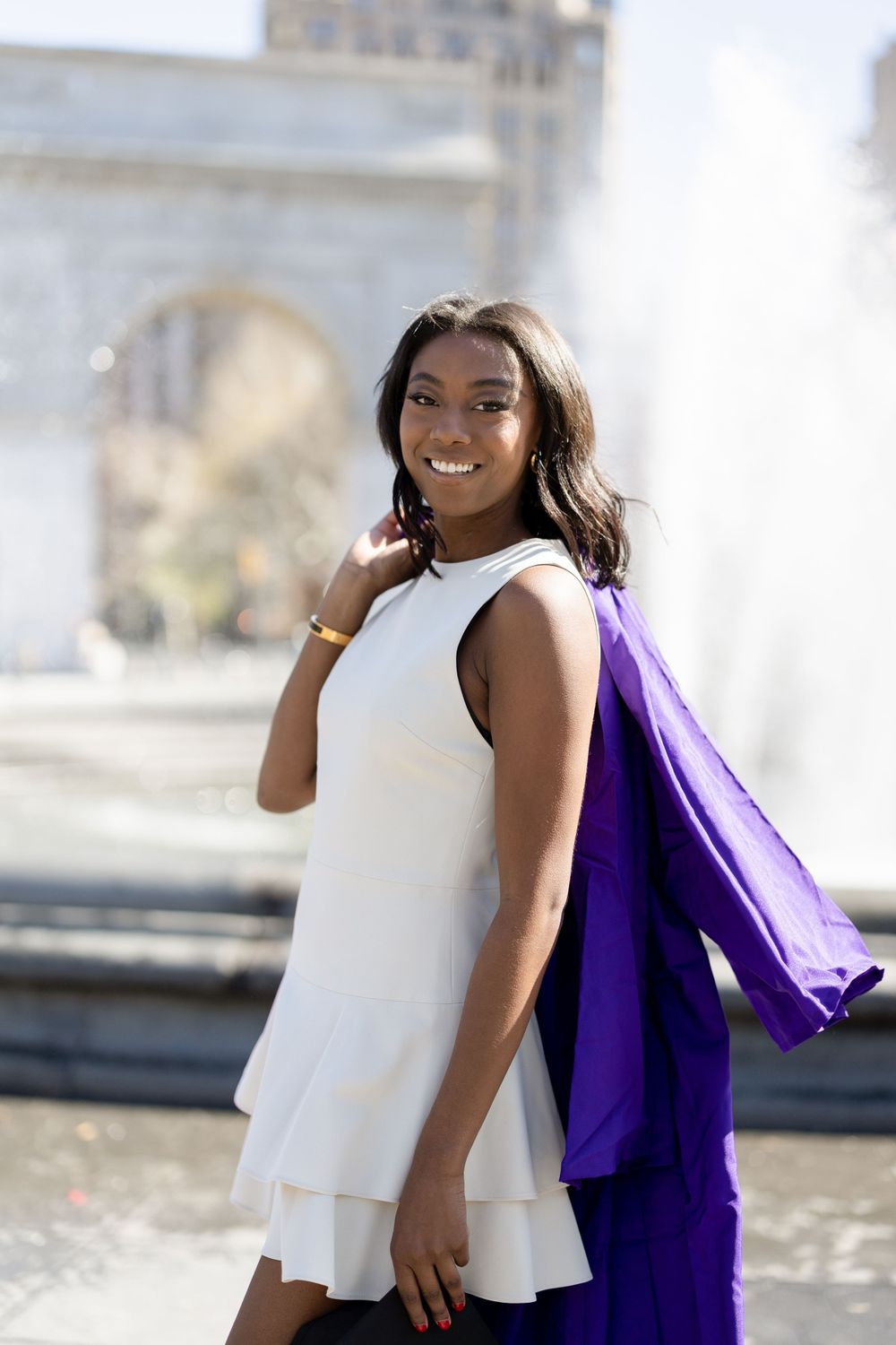 Graduate in white dress with purple graduation gown poses by fountain in Washington Square Park.