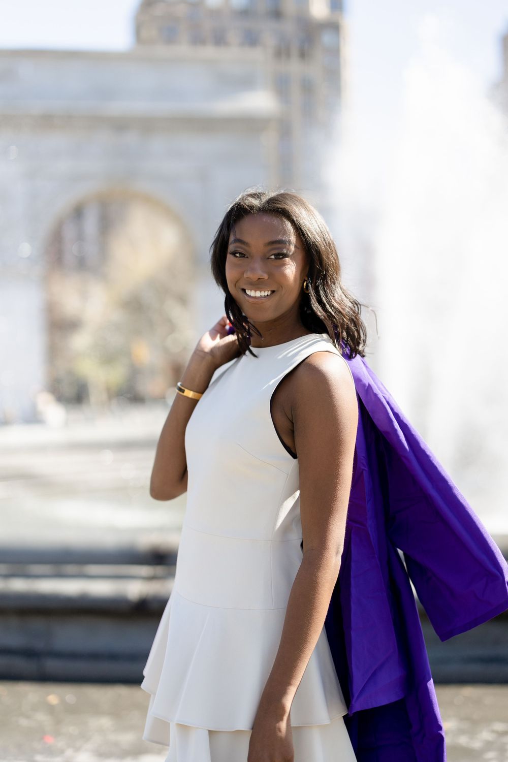 Graduate in white dress and purple gown smiles while posing near Washington Square Arch fountain.