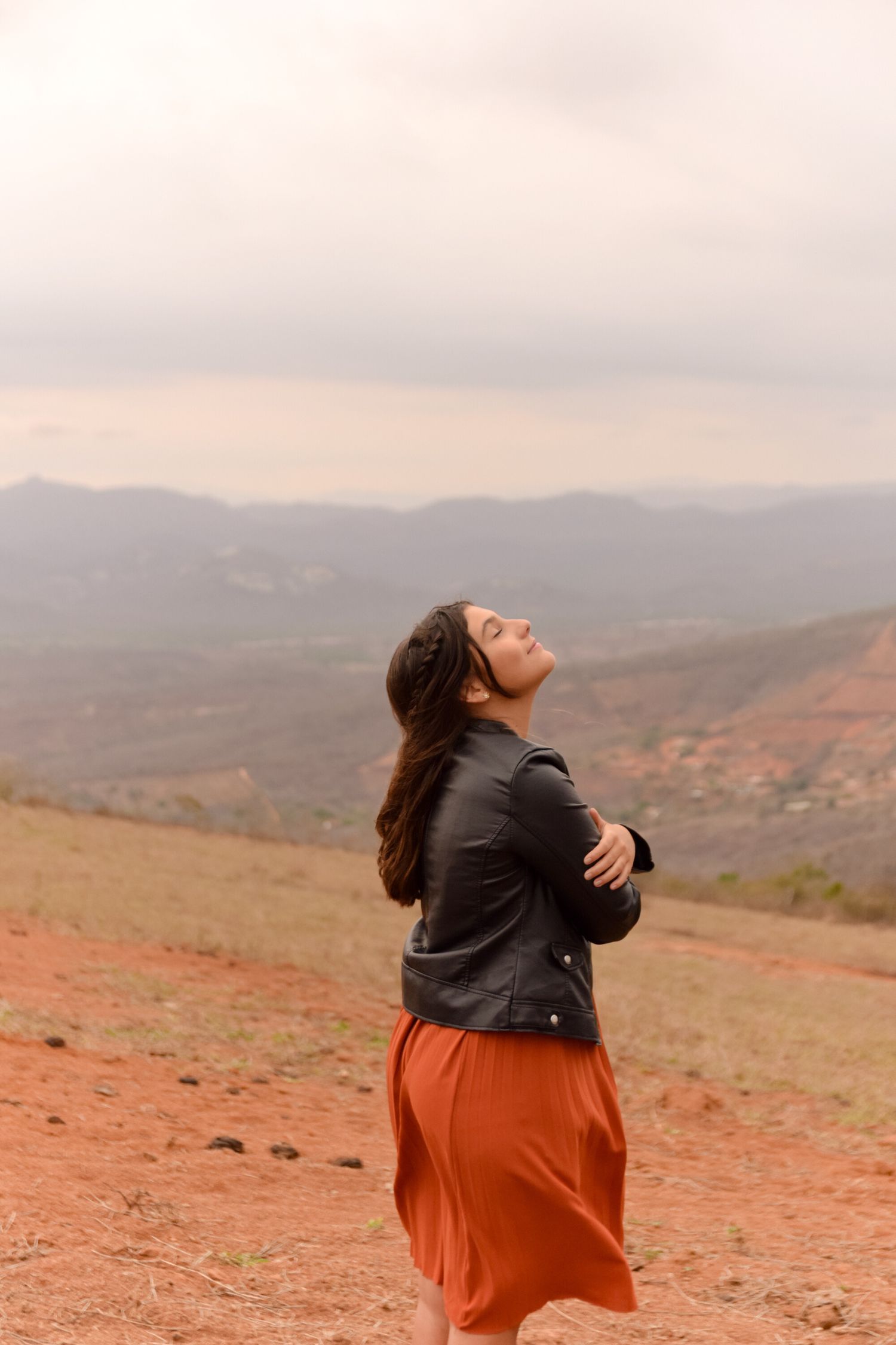 A person in a leather jacket and orange skirt stands on a hilltop overlooking mountains at sunset.