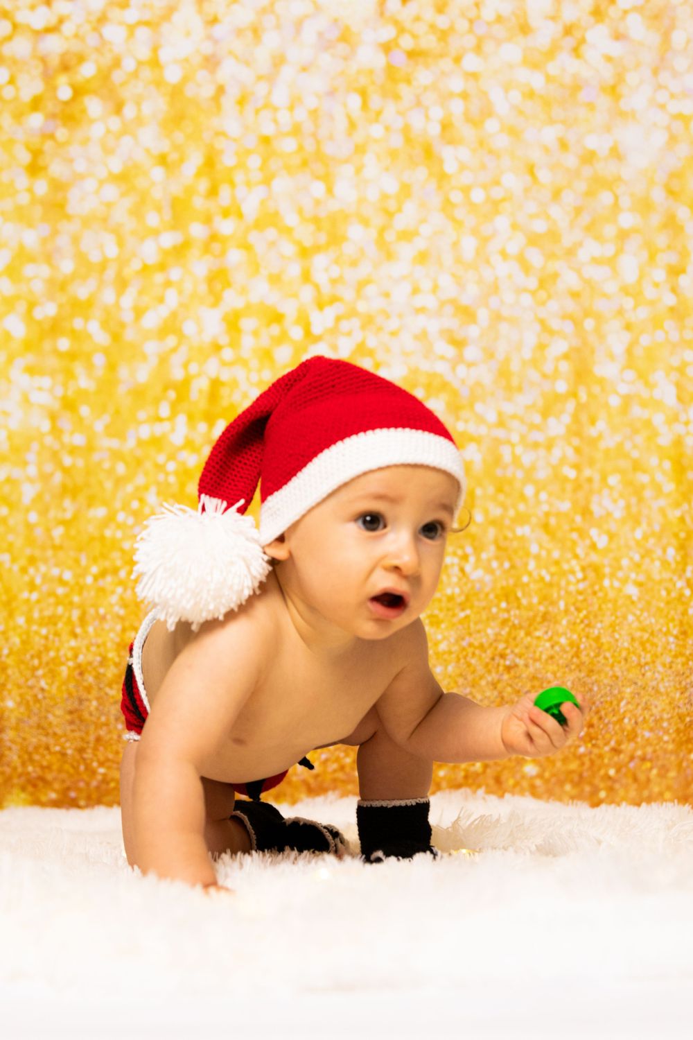 Baby wearing Santa hat crawling on white surface against golden sparkly background.