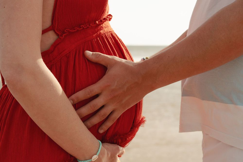Close up of hands tenderly touching pregnant belly in red dress on beach.