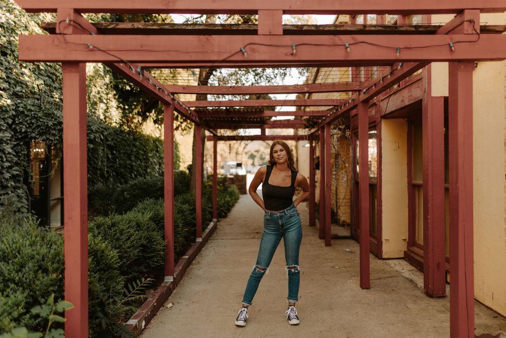 Person in black top and jeans poses under rustic wooden pergola walkway during golden hour.