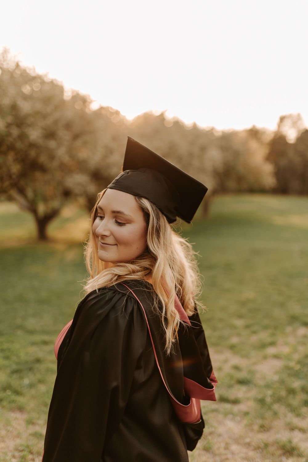 A graduate in traditional academic regalia poses during sunset in an orchard.