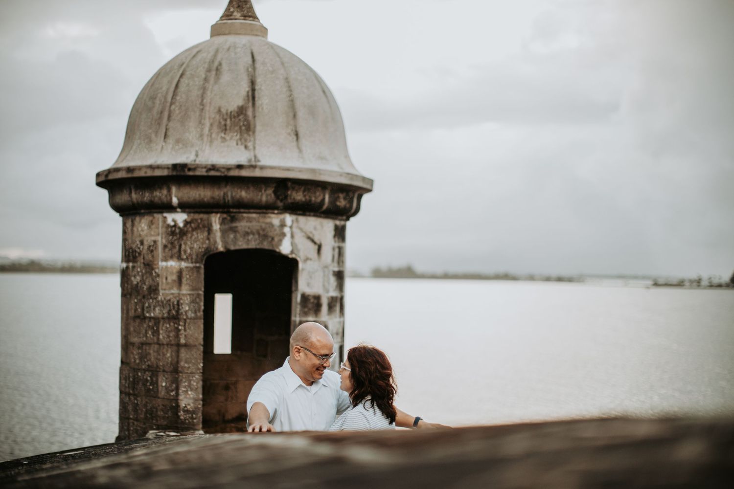 Couple sharing a romantic moment by a historic stone lookout tower overlooking the water.